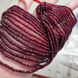 Puede incluir: Hebras de cuentas de piedras preciosas facetadas de color rojo intenso. Las cuentas son pequeñas y de tamaño uniforme, ensartadas para crear múltiples hebras. El color es un rojo oscuro intenso, con un ligero brillo de las facetas. Las cuentas se sostienen en una mano.