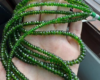 May include: A strand of green gemstone beads, likely peridot, with a faceted, slightly rounded shape. The beads are strung on a clear thread and are arranged in a loose, flowing pattern.