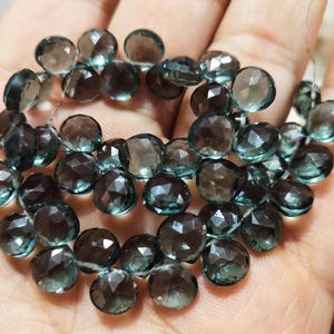 May include: A collection of faceted, round gemstones in shades of teal and gray. The gemstones are strung together, showcasing their polished surfaces and intricate cuts. The beads are held in a hand, highlighting their size and color.