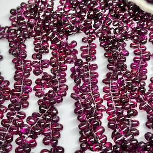 Könnte beinhalten: Nahaufnahme einer Kette aus tiefvioletten Edelsteinperlen. Die Perlen sind klein, tropfenförmig und mit weißem Faden aufgefädelt. Die Perlen haben eine glänzende Oberfläche und sind in vertikalen Reihen angeordnet.