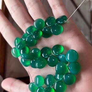 Op de afbeelding: Een streng gepolijste, ovaalvormige groene kralen. De kralen zijn geregen met een dunne, zilverkleurige draad. De kralen zijn een levendige, doorschijnende groene kleur en hebben een gladde, glanzende afwerking. De kralen worden in een hand gehouden. Elke kraal is ongeveer 1 cm in diameter.