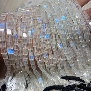 Könnte beinhalten: Stränge aus klaren, rechteckigen Perlen mit schillernden blauen und weißen Reflexen. Die Perlen sind aufgefädelt, wodurch ein schimmernder Effekt entsteht. Die Stränge werden in einer Hand gehalten, mit schwarzen und weißen Quasten am unteren Ende. Ideal für die Schmuckherstellung.