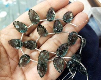 Puede incluir: Primer plano de piedras preciosas en forma de hoja, en tonos turquesa y gris, ensartadas en alambre de plata. Las cuentas facetadas tienen una superficie detallada y texturizada. Las piedras están dispuestas en un patrón repetitivo.
