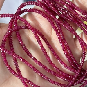 May include: Strands of faceted, deep red ruby beads. The beads are small and strung together, creating a delicate and shimmering effect. The ruby beads are held in a hand, showcasing their rich color and texture. Ideal for jewelry making.