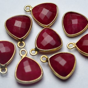 Peut inclure: Gros plan sur huit pendentifs triangulaires en pierres précieuses rouge rubis facettées, avec des montures dorées et de petites boucles pour la fixation de bijoux. Les pendentifs sont disposés sur une surface blanche, mettant en valeur leur couleur riche et leur design.