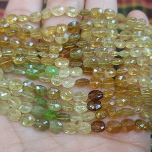 May include: A collection of oval-shaped, faceted gemstones in yellow, green, and brown hues. The beads are strung together, forming multiple strands. The gemstones have a polished surface, reflecting light.