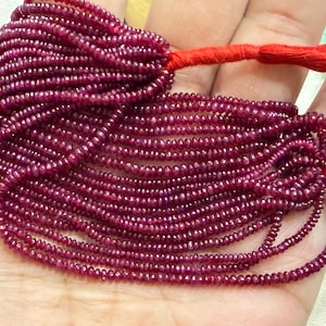 Op de afbeelding: Een meerstrengs ketting van kleine, gefacetteerde, diep magenta gekleurde robijnkralen. De strengen zijn samengevoegd en aan de bovenkant vastgezet met een felrode draad, wat zorgt voor een dichte, gelaagde uitstraling van gepolijste, ronde edelsteenkralen.