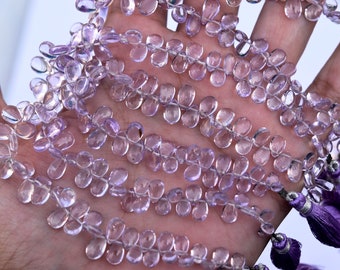 May include: A strand of pale lavender gemstone beads, each bead is faceted and shaped like a teardrop. The beads are strung on a thin wire and are approximately 10mm in diameter. The beads are displayed on a hand, with the ends of the strand hanging down.