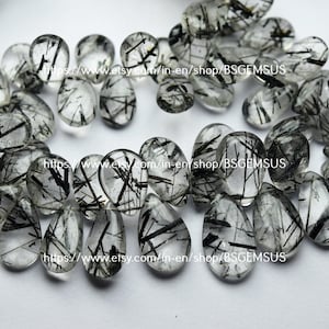 May include: A collection of 20 teardrop-shaped, clear quartz stones with black tourmaline inclusions. The stones are approximately 1 inch long and have a smooth, polished finish.