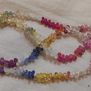 Könnte beinhalten: Eine Strähne aus bunten Edelsteinperlen, die einen Regenbogen aus Farben wie Rosa, Blau, Gelb und Grün aufweist. Die Perlen sind facettiert und haben eine birnenförmige Form.