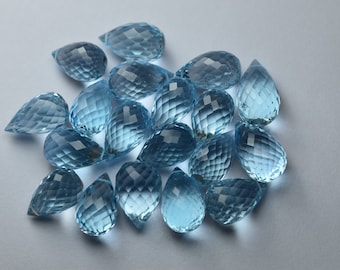 Puede incluir: Un conjunto de gemas azules en forma de lágrima. Las piedras están facetadas, lo que les da un aspecto brillante. Son de color azul claro y están dispuestas sobre una superficie blanca. Cada gema tiene un extremo puntiagudo.