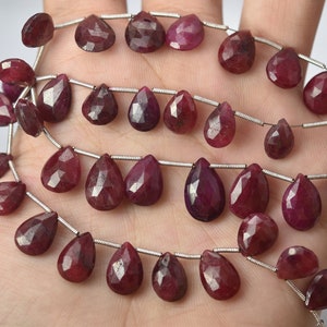 May include: A strand of ruby gemstone beads in a teardrop shape. The beads are strung on a silver wire and are a deep red color.
