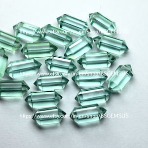 Puede incluir: Un grupo de 17 formas de cristal facetadas, de color verde claro, con doble punta. Los cristales están dispuestos en un montón suelto sobre una superficie blanca.