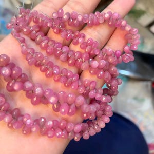 Op de afbeelding: Strengen roze edelsteen kralen, elk in een druppelvorm, zijn aan elkaar geregen. De kralen zijn felroze en hebben een facetgeslepen oppervlak dat het licht vangt. De kralen worden in een hand gehouden, waardoor hun kleur en vorm worden benadrukt.