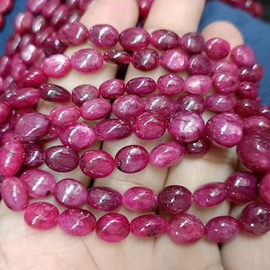 May include: A strand of ruby red gemstone beads, each bead is oval shaped and has a smooth, polished finish. The beads are strung together on a clear thread and are displayed on a hand.