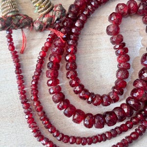 May include: A close-up of a multi-strand necklace featuring faceted, deep red gemstones. The necklace has an ornate clasp with gold-colored metal and decorative fabric accents. The gemstones are arranged in graduated sizes.