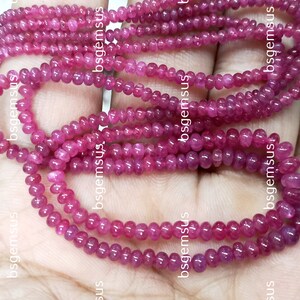 May include: A strand of pink ruby beads, approximately 3mm in diameter, with a smooth, polished finish. The beads are strung on a clear thread and are arranged in a loose, flowing pattern.