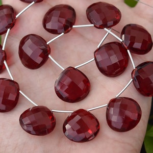 May include: Strand of faceted, heart-shaped, deep red gemstone beads. The beads are strung on a silver-colored wire. The beads are held in a hand, with a dark background and green foliage visible.