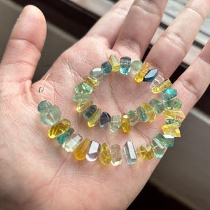 Puede incluir: Un collar o pulsera de cuentas de piedras preciosas multicolores. Las cuentas son de color amarillo, verde azulado y transparente. Las cuentas de forma irregular están ensartadas. Las piedras preciosas se sostienen en una mano.