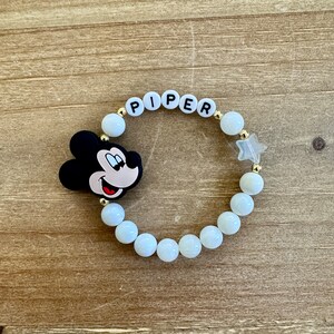 Puede incluir: Pulsera de cuentas blancas con un dije de Mickey Mouse, cuentas doradas y el nombre "PIPER" escrito en letras blancas.