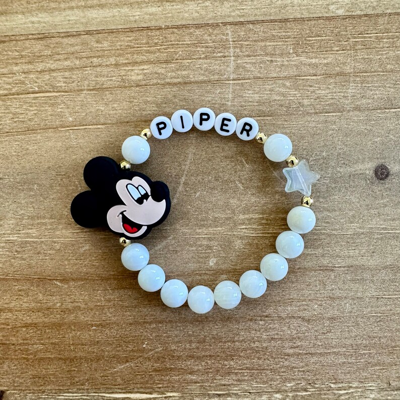 Puede incluir: Pulsera de cuentas blancas con un dije de Mickey Mouse, cuentas doradas y el nombre "PIPER" escrito en letras blancas.