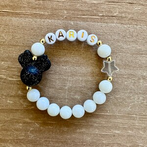 May include: White beaded bracelet with gold letters spelling "KARIS" and a black and white glitter Mickey Mouse charm. The bracelet also features a clear star charm.