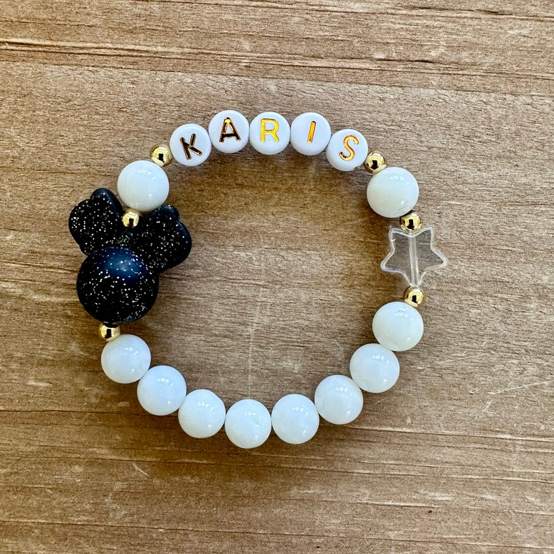 May include: White beaded bracelet with gold letters spelling "KARIS" and a black and white glitter Mickey Mouse charm. The bracelet also features a clear star charm.