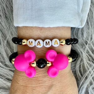 May include: Two bracelets on a wrist. The top bracelet has black beads with gold accents and white letter beads spelling "MAMA". The bottom bracelet has black beads with gold accents and pink Mickey Mouse head charms.