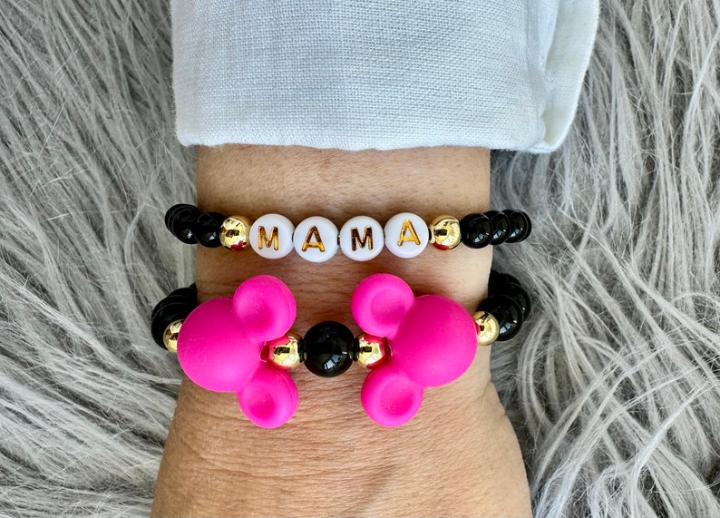 May include: Two bracelets on a wrist. The top bracelet has black beads with gold accents and white letter beads spelling "MAMA". The bottom bracelet has black beads with gold accents and pink Mickey Mouse head charms.