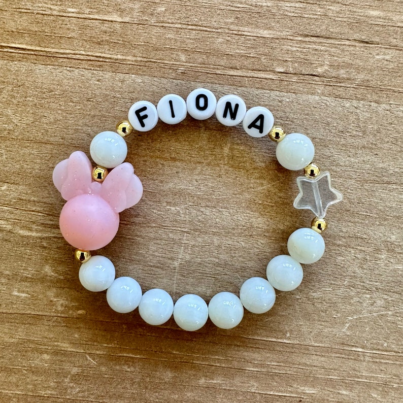 May include: A white beaded bracelet with gold spacers, featuring the name "Fiona" spelled out in black letters. The bracelet also includes a pink bow charm and a clear star charm.