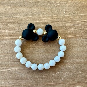 May include: A white beaded bracelet with black Mickey Mouse charms. The bracelet is made with white beads and gold accents.