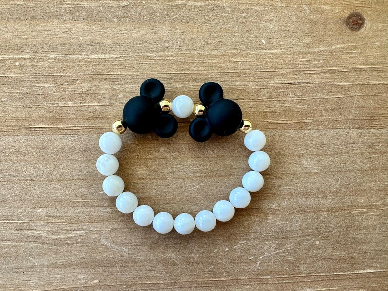 May include: A white beaded bracelet with black Mickey Mouse charms. The bracelet is made with white beads and gold accents.