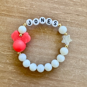 May include: A white beaded bracelet with gold spacers and a red reindeer charm. The bracelet also has white letter beads spelling out the name "JONES".