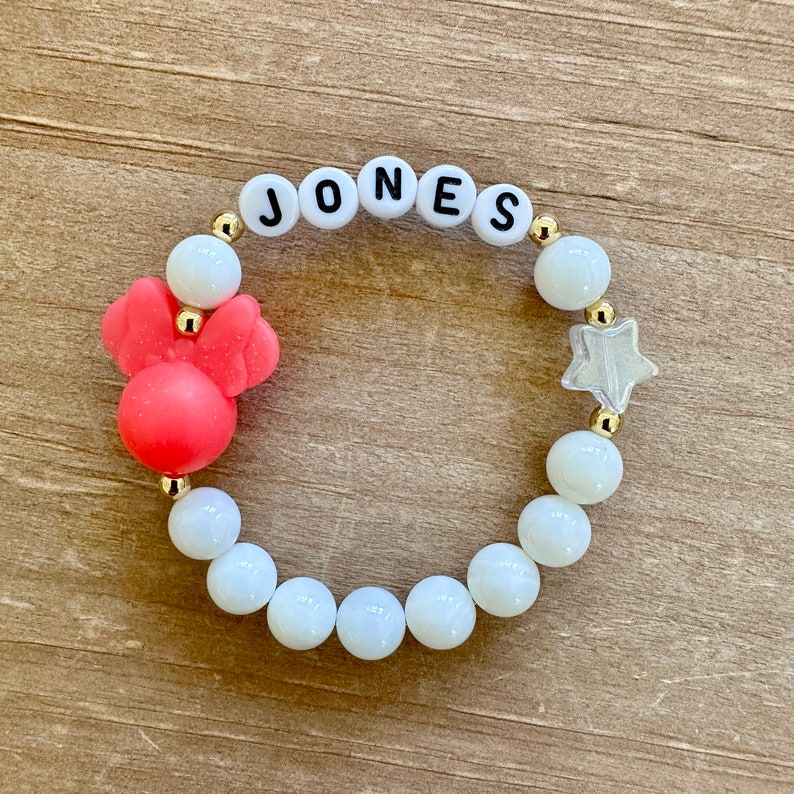 May include: A white beaded bracelet with gold spacers and a red reindeer charm. The bracelet also has white letter beads spelling out the name "JONES".