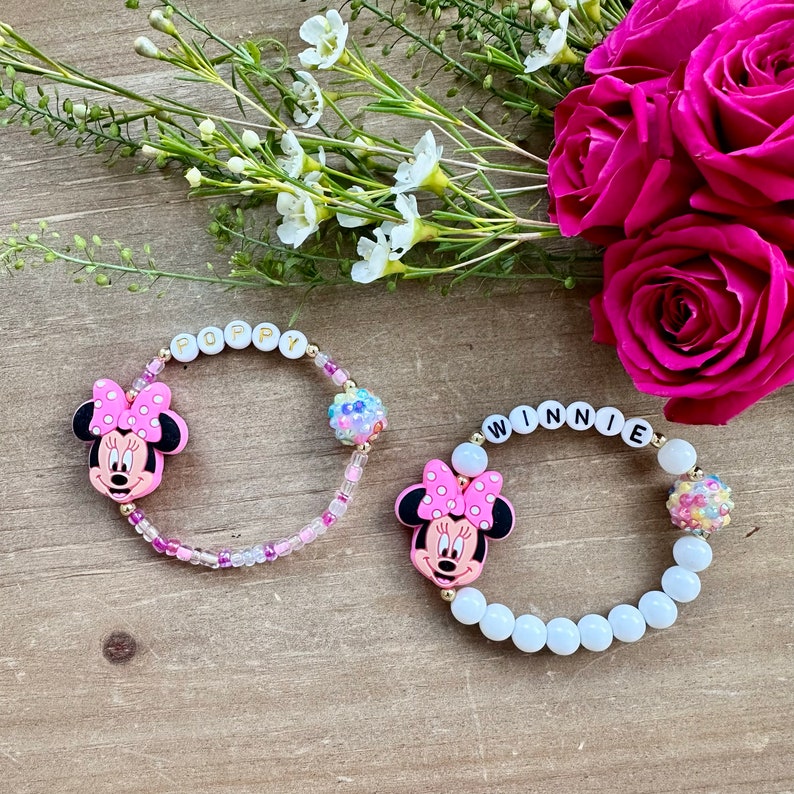 May include: Two beaded bracelets with Minnie Mouse charms. One bracelet is white with the name "WINNIE" spelled out in black letters. The other bracelet is pink and white with the name "POPPY" spelled out in white letters.