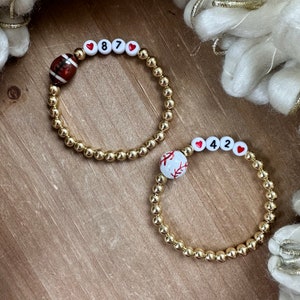 May include: Two gold beaded bracelets with charms. One bracelet has a football charm and the numbers 87 with red hearts on either side. The other bracelet has a baseball charm and the numbers 42 with red hearts on either side.