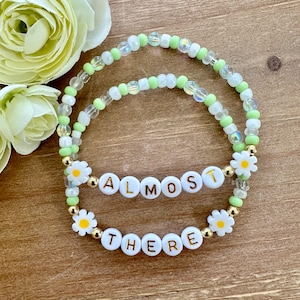 May include: Two beaded bracelets with green and white beads. The bracelets have white letter beads that spell out "ALMOST" and "THERE" with a daisy charm on each bracelet.