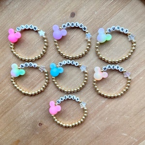 May include: Seven gold bead bracelets with a Mickey Mouse charm and a star charm. Each bracelet has a different name spelled out on it: Sloan, Harlow, Sunny, Addi, Riley, Maeve, and Everly.