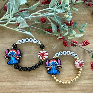 Puede incluir: Dos pulseras de cuentas con los nombres "GEORGE" y "PARKER". Cada pulsera presenta un dije de personaje de dibujos animados azul sosteniendo un bastón de caramelo y una cuenta de menta piperita roja y blanca. Una pulsera es negra, la otra dorada.