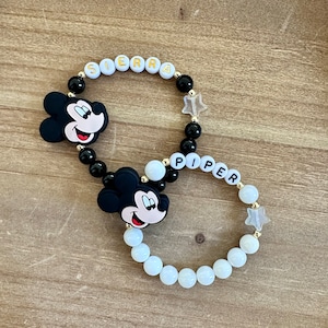 Puede incluir: Dos pulseras de cuentas blancas y negras con dijes de Mickey Mouse y cuentas de letras doradas que deletrean los nombres "SIERRA" y "PIPER".