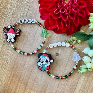 May include: Two beaded bracelets with the names "EMMA" and "ROSIE" spelled out in white letter beads. Each bracelet features a Mickey and Minnie Mouse Christmas charm, along with Christmas tree charms and colorful beads.
