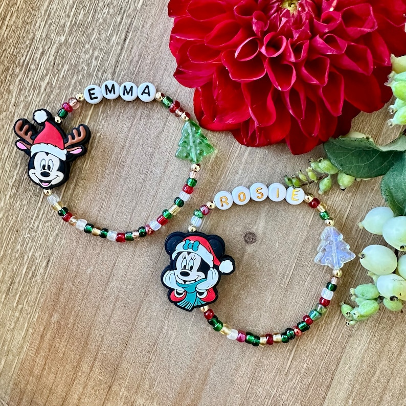 May include: Two beaded bracelets with the names "EMMA" and "ROSIE" spelled out in white letter beads. Each bracelet features a Mickey and Minnie Mouse Christmas charm, along with Christmas tree charms and colorful beads.