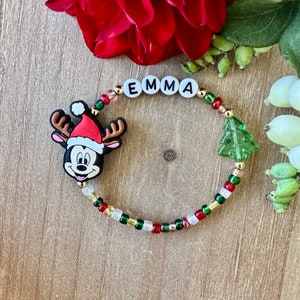 May include: A beaded bracelet with the name "EMMA" spelled out in white letter beads. The bracelet features a Mickey Mouse charm wearing a Santa hat and a green Christmas tree charm. The beads are red, green, gold, and clear.