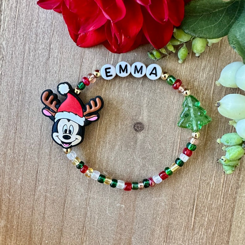 May include: A beaded bracelet with the name "EMMA" spelled out in white letter beads. The bracelet features a Mickey Mouse charm wearing a Santa hat and a green Christmas tree charm. The beads are red, green, gold, and clear.