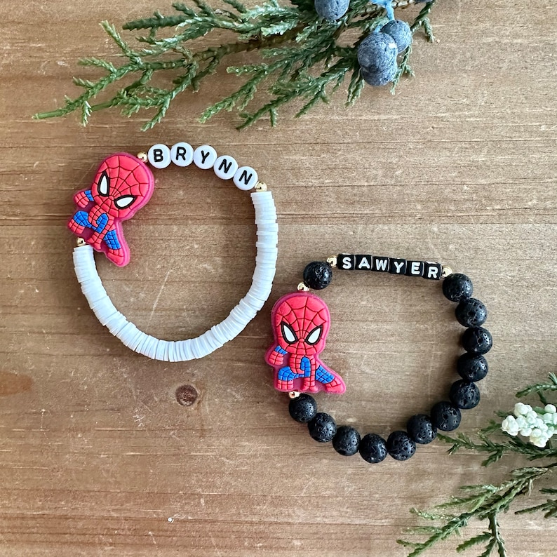 May include: Two bracelets with Spiderman charms. One bracelet is white with the name "BRYNN" spelled out in black letters. The other bracelet is black with the name "SAWYER" spelled out in white letters.