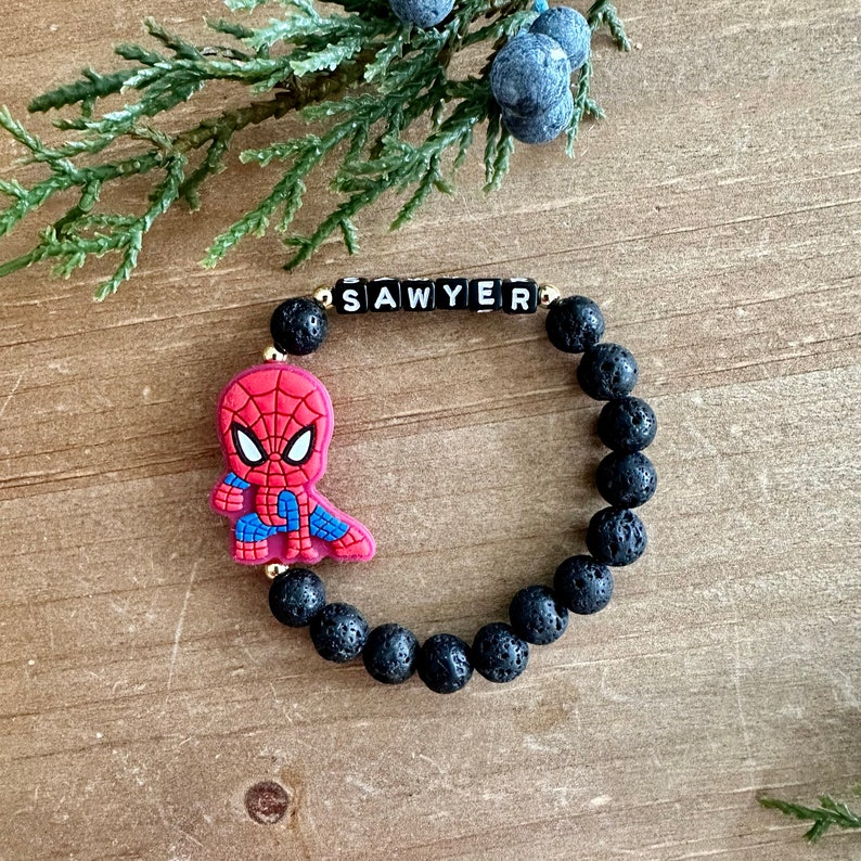 May include: Black lava rock bracelet with a gold bead and a small red and blue Spiderman charm. The bracelet has the name "SAWYER" spelled out in black letters on gold beads.