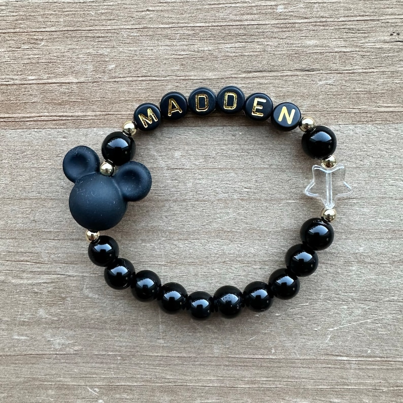 May include: Black beaded bracelet with a Mickey Mouse charm, a clear star charm, and gold letter beads spelling out the name "MADDEN".