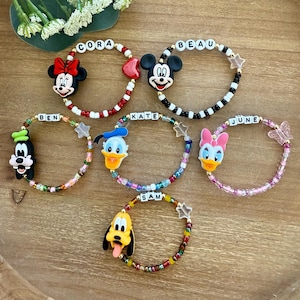 May include: A collection of beaded bracelets featuring Disney character charms. Each bracelet has a different character, including Mickey Mouse, Minnie Mouse, Donald Duck, Daisy Duck, Goofy, and Pluto, with a name spelled out in letter beads.