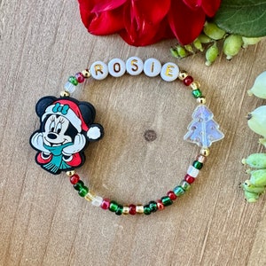 May include: A beaded bracelet with the name "ROSIE" spelled out in white letter beads. The bracelet features a Minnie Mouse charm in a Santa hat and a Christmas tree charm. The beads are red, green, gold, and white.