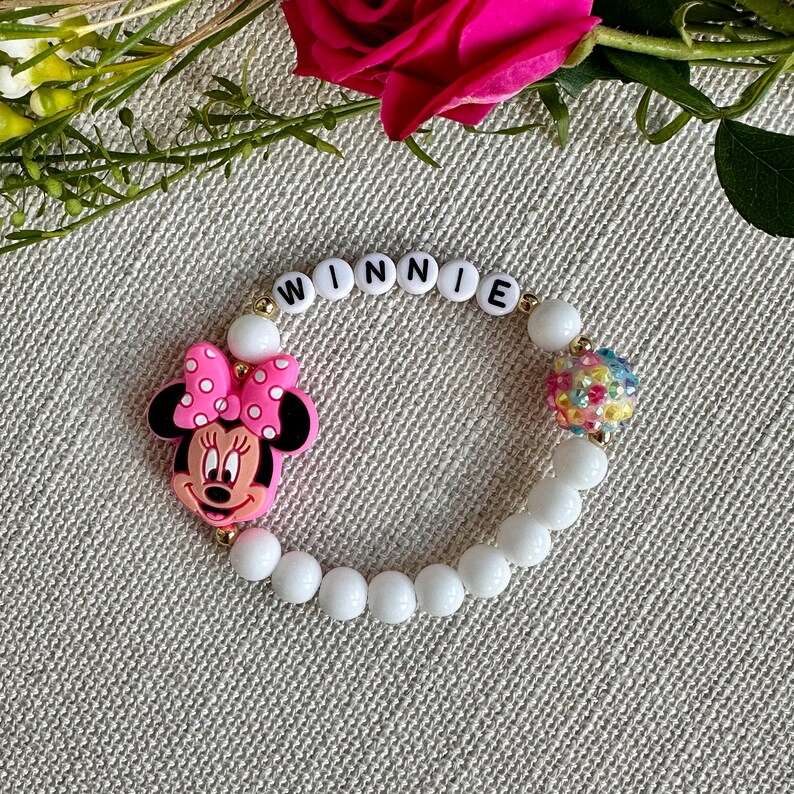 May include: White beaded bracelet with the name "WINNIE" spelled out in black letters. The bracelet also features a pink Minnie Mouse charm and a colorful, sparkly bead.